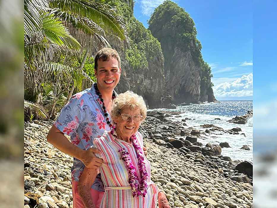An older woman and a younger man pose on a rocky beach with palm leaves behind them.