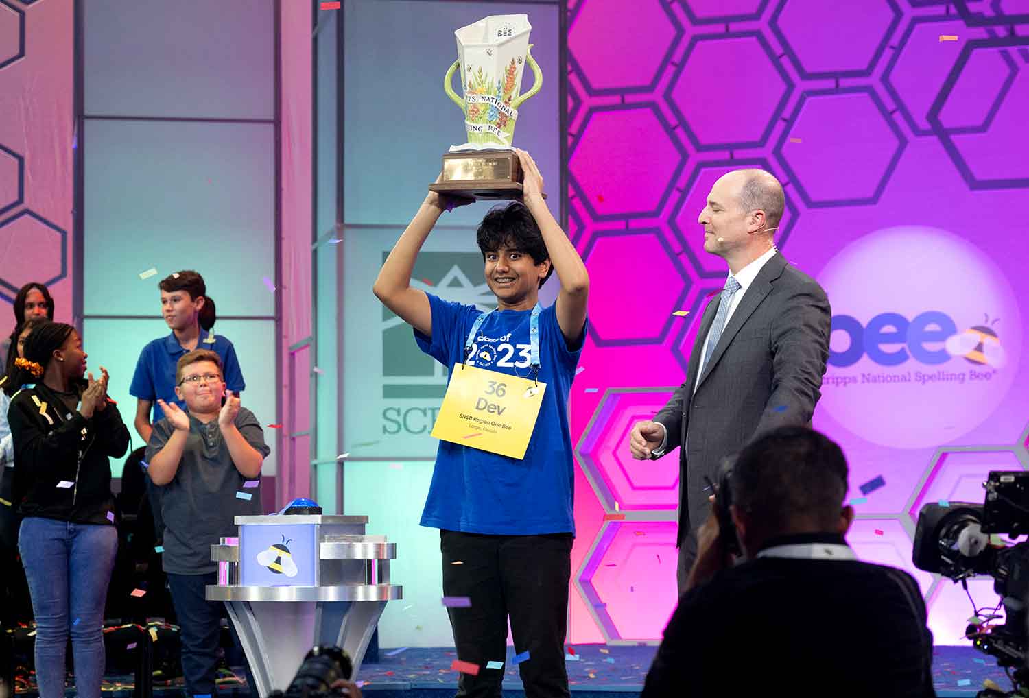 A teen on a stage smiles and holds up a trophy as an audience and other teens clap.