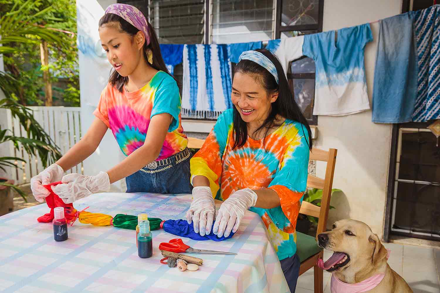 A woman and a girl dying a rolled and banded shirt with tie dyed clothing hanging in the background.