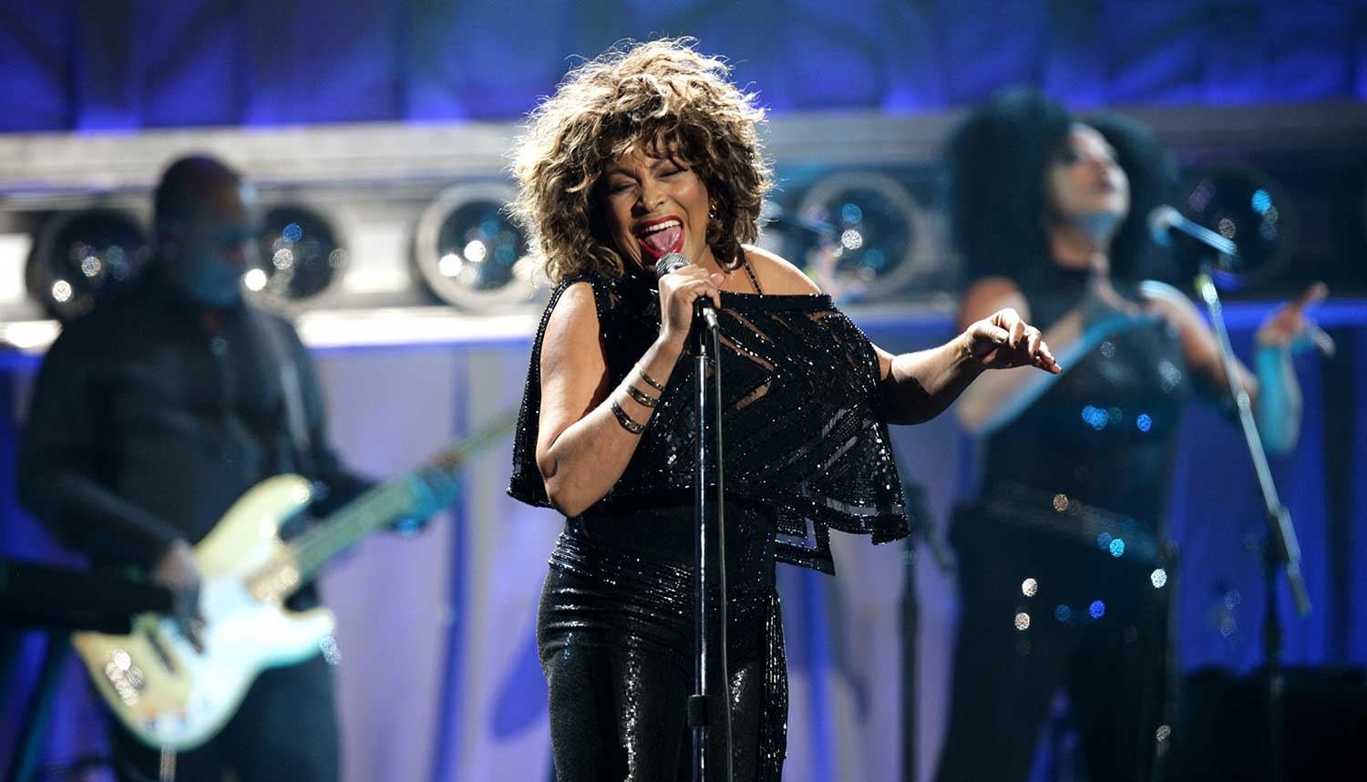 Tina Turner sings into a microphone with a backup singer and a guitar player in the background.