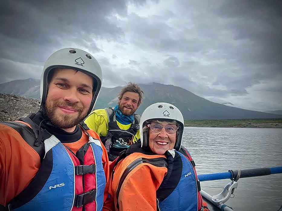 An older woman and two younger men wearing rafting gear pose near a body of water with a mountain behind them.