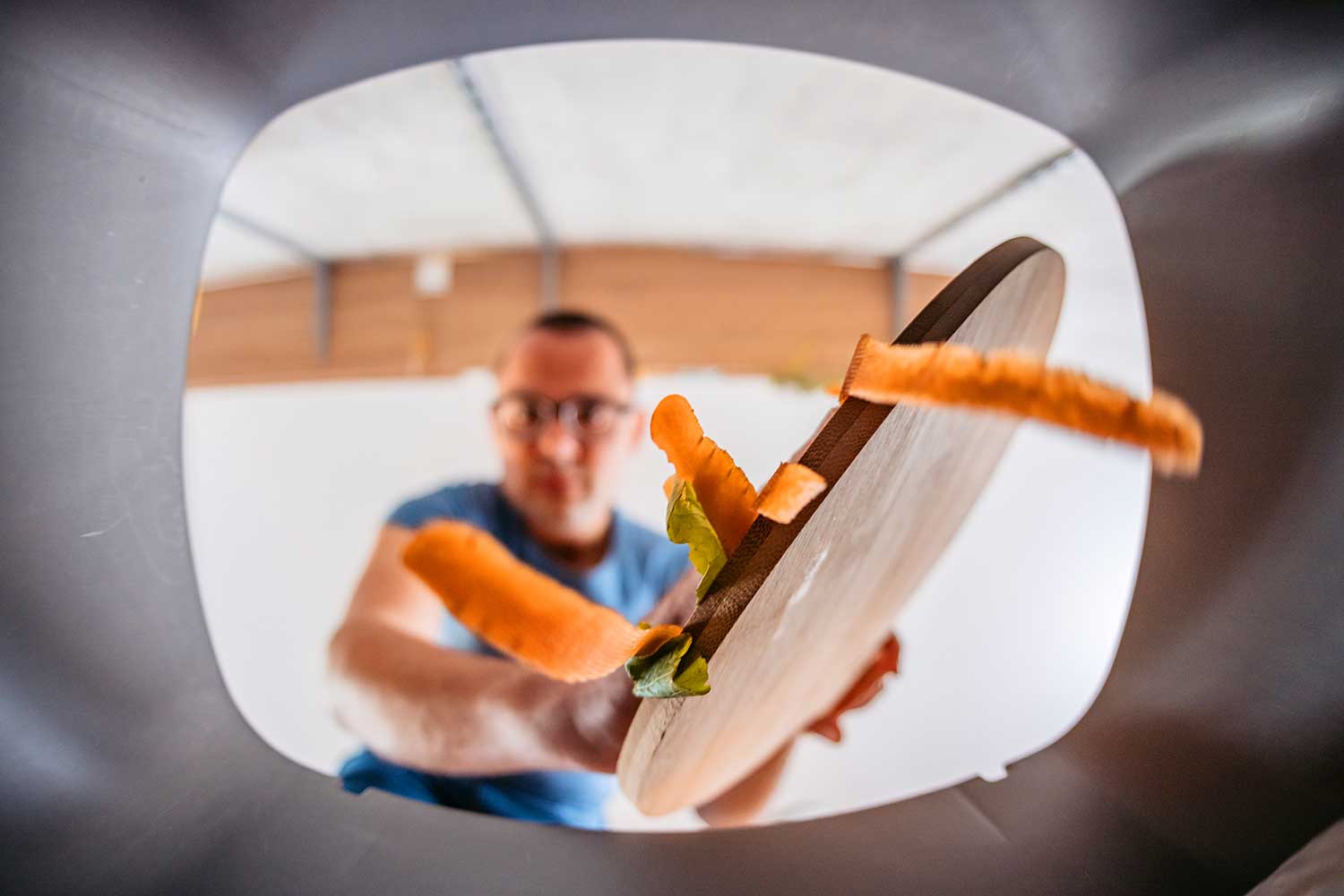 A man scrapes carrots and lettuce from a cutting board to a container as seen from inside the container.