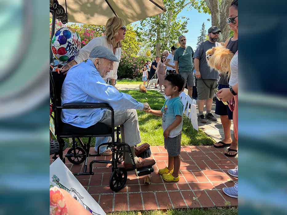 A seated older man shakes hands with a young child while a group of people and dogs are lined up behind the child.