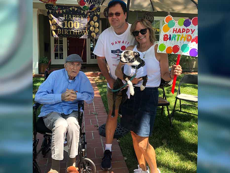 Two people stand next to a seated older man and pose for a photo. One of them holds a small dog and a happy birthday sign.