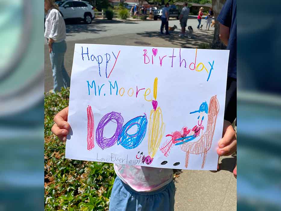 An unseen child holds up a drawing that says happy birthday Mr Moore and shows a man with a dog on his lap.