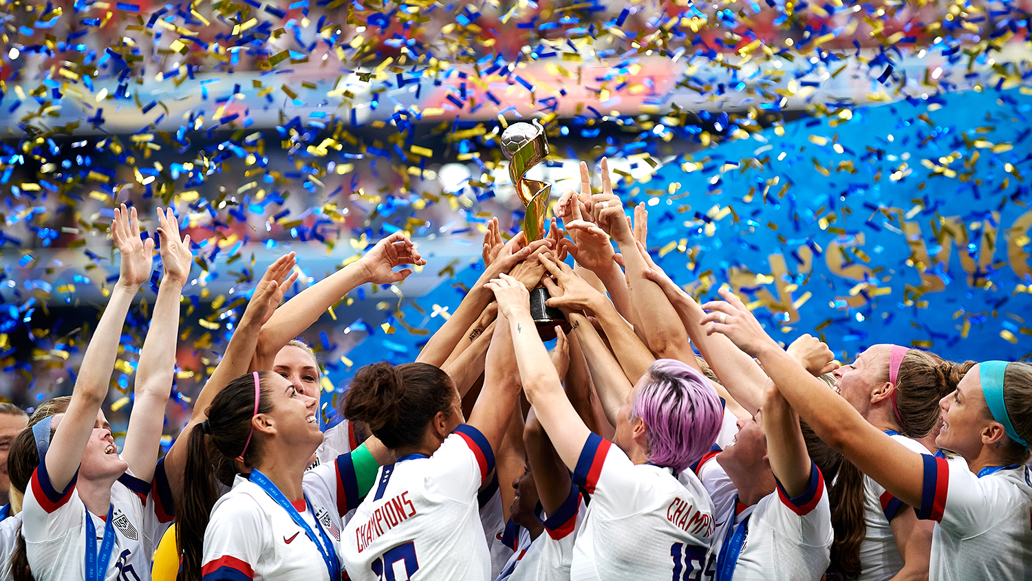 A group of uniformed soccer players gathered and reaching up to touch a trophy as confetti falls.