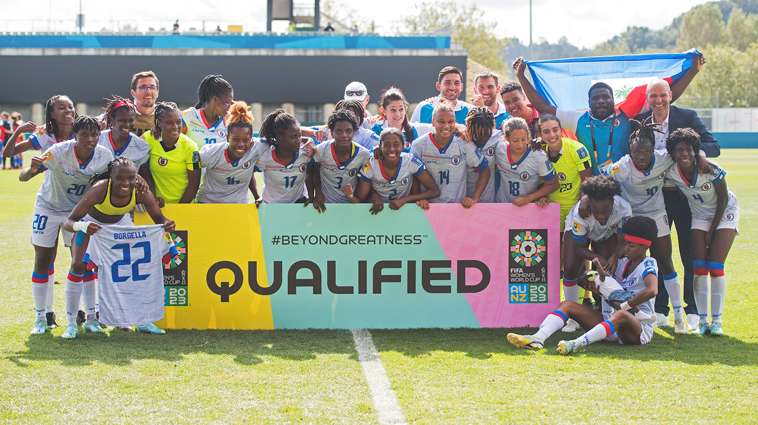 A team of women in uniforms and some men pose on a soccer field with a banner that says qualified.