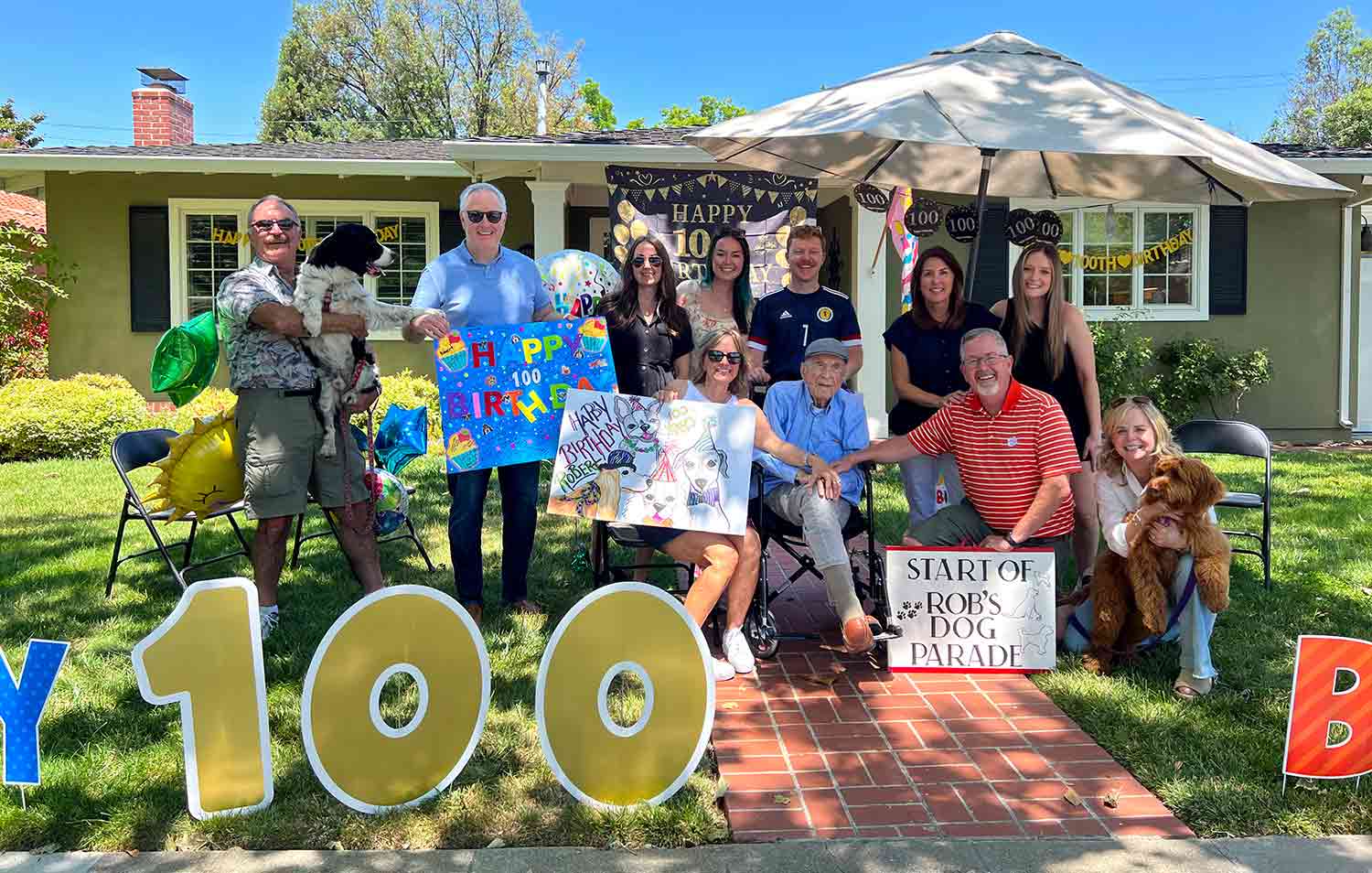 A group of people and two dogs pose in front of a home with birthday decorations, happy birthday signs, and the number 100.