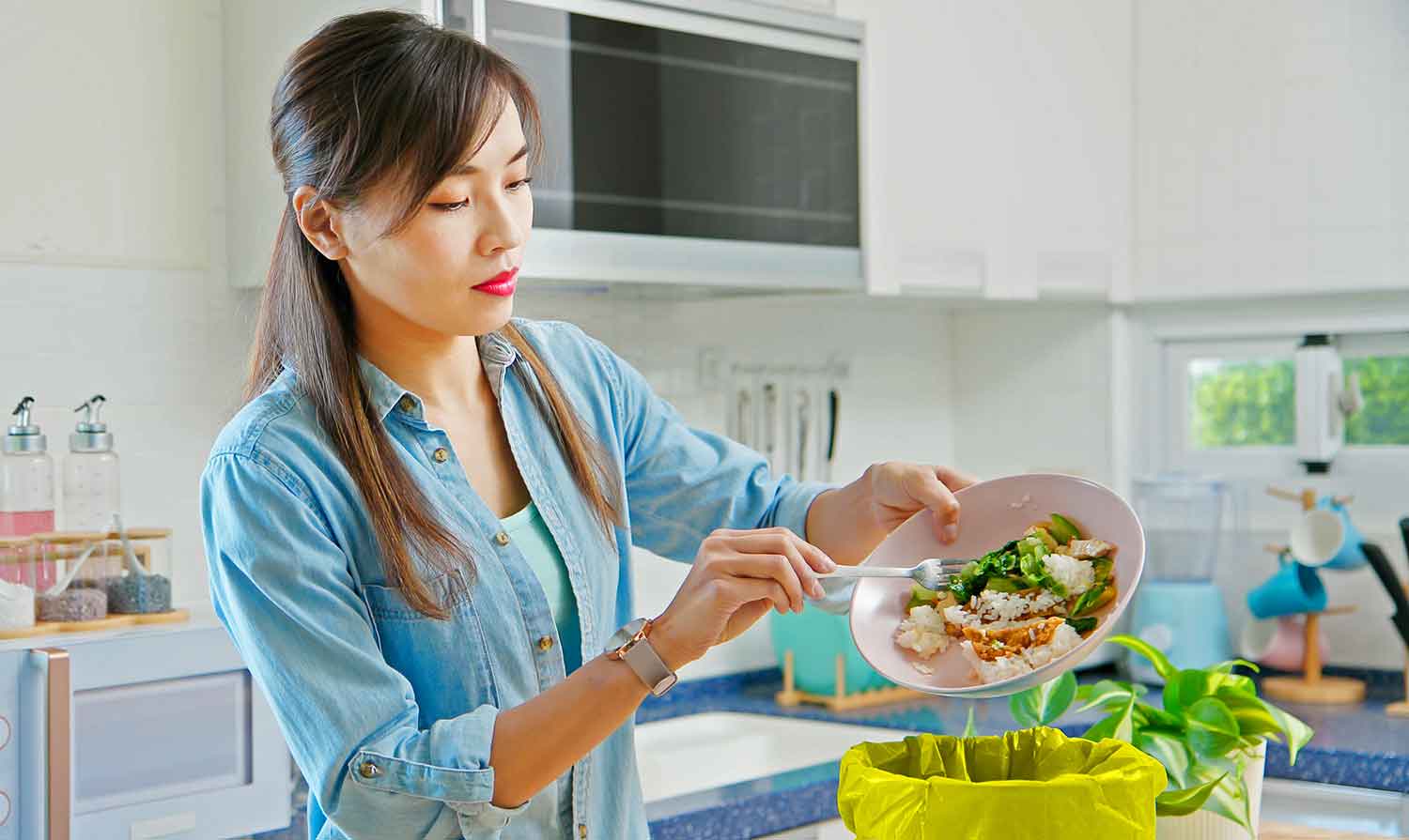 A woman in a kitchen transfers food from a bowl to a bin lined with yellow plastic.