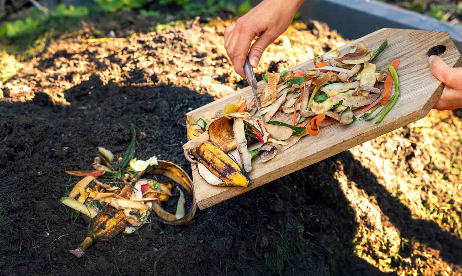 A knife is used to scrap food scraps from a wooden cutting board to a compost pile.
