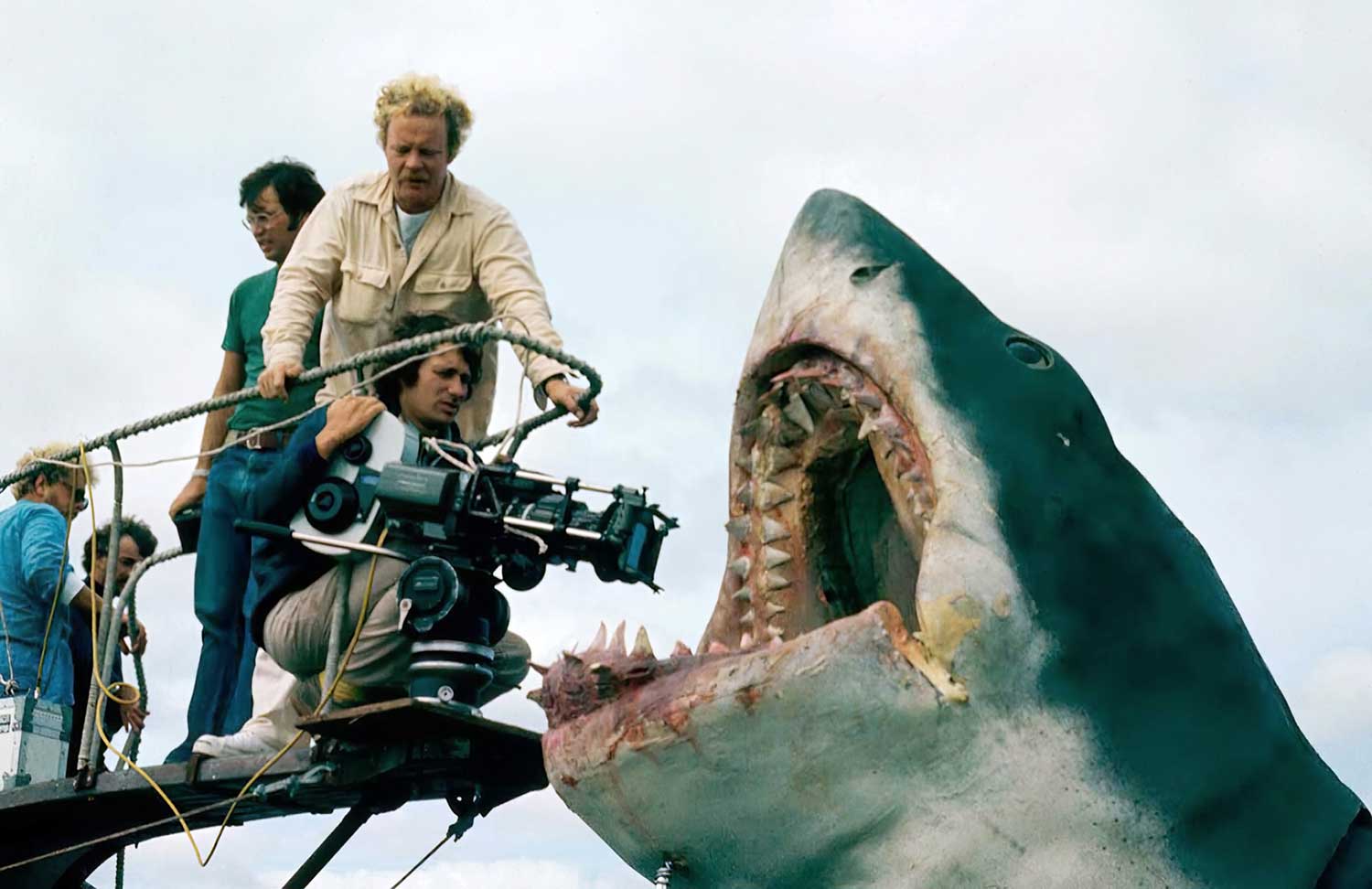 Steven Spieberg crouches on a crane pointing a camera into the mouth of a fake shark as four other men stand behind him.