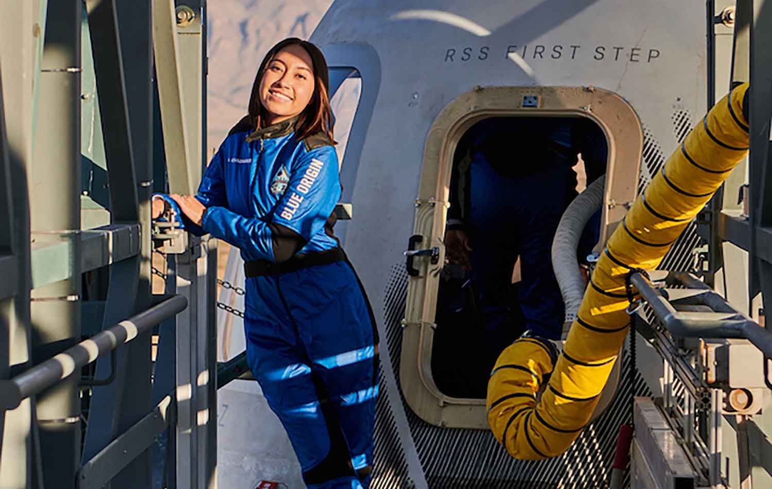 A woman in a blue Blue Origin space suit poses outside of a space capsule.