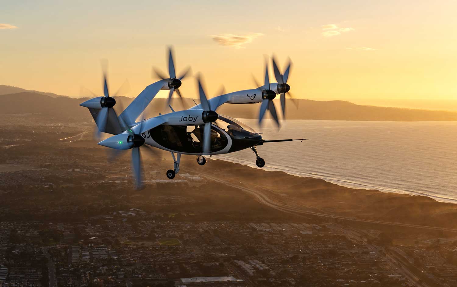 A vehicle with six vertical propellers in flight over a coastline.
