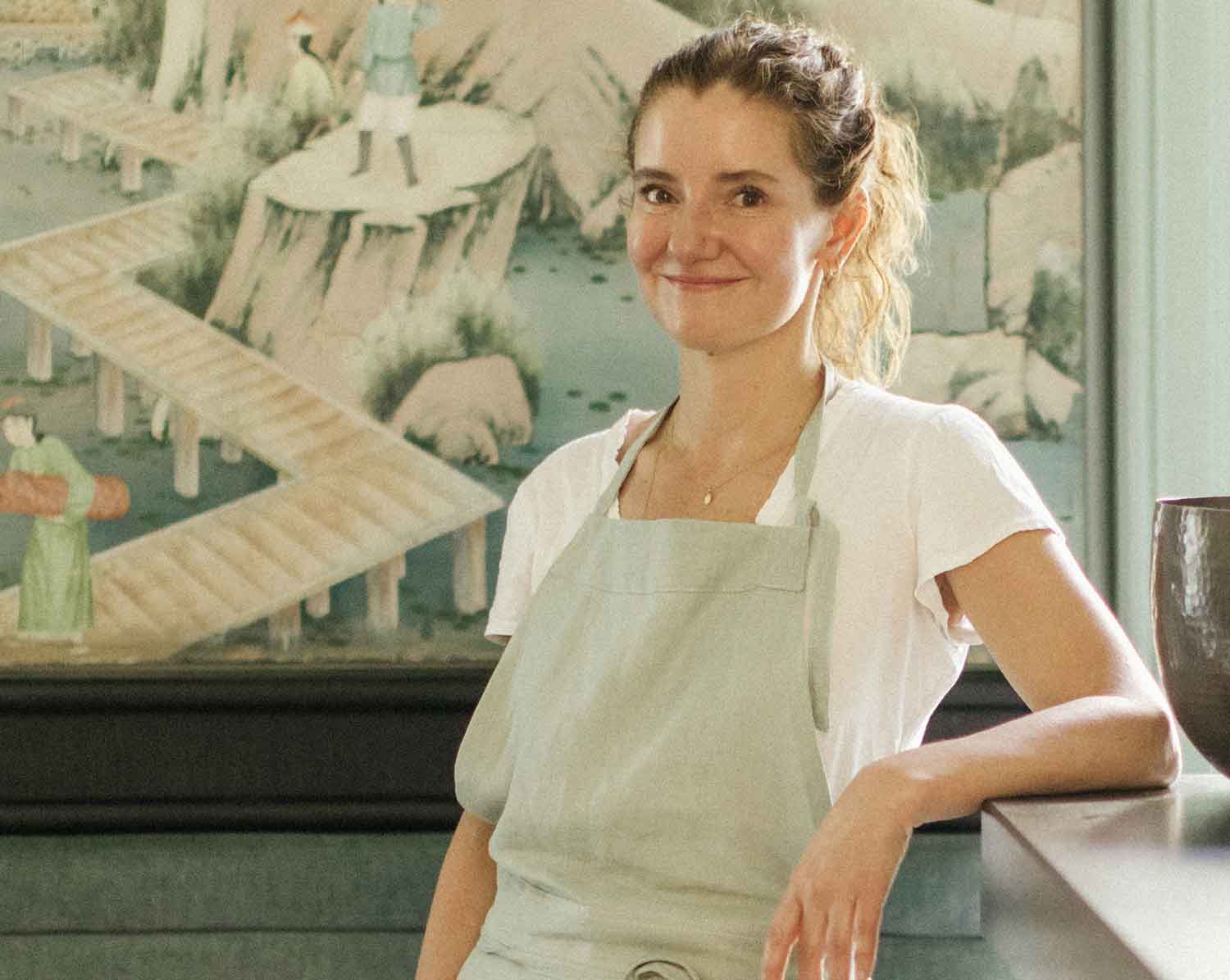 A woman in an apron poses in front of a painting.