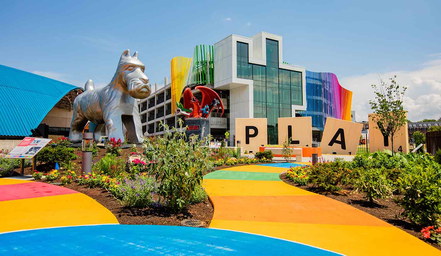 Giant Scrabble tiles spell the word play next to a giant Monopoly dog among colorful paths and plantings outside of a building.