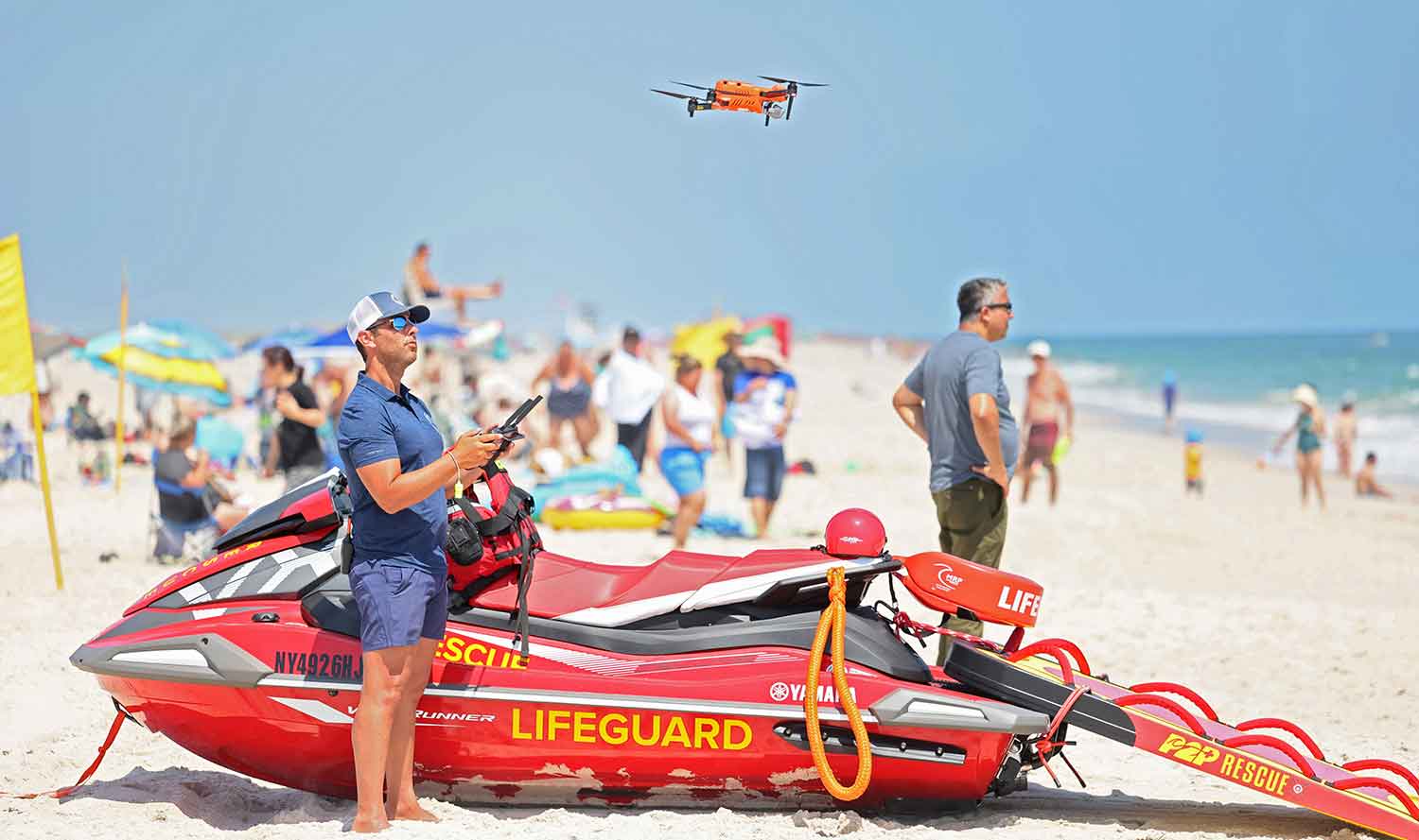 A man on a beach stands in front of a red jet ski that says Lifeguard as he operates a drone that is flying nearby.