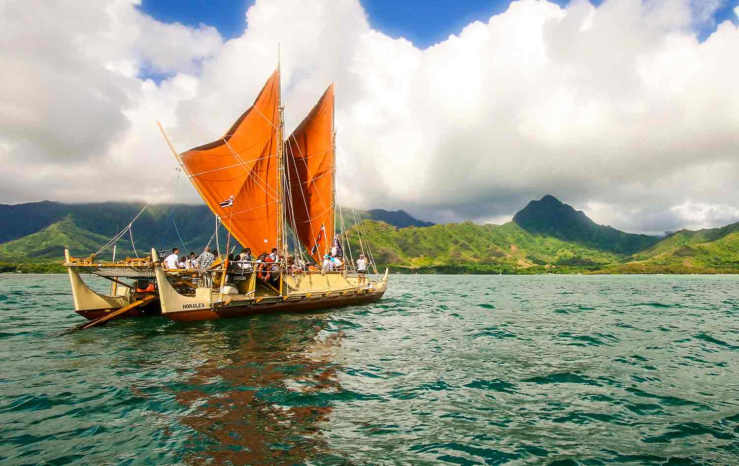 A replica of an ancient canoe with orange sails carries several people on a body of water with green mountains in the background.