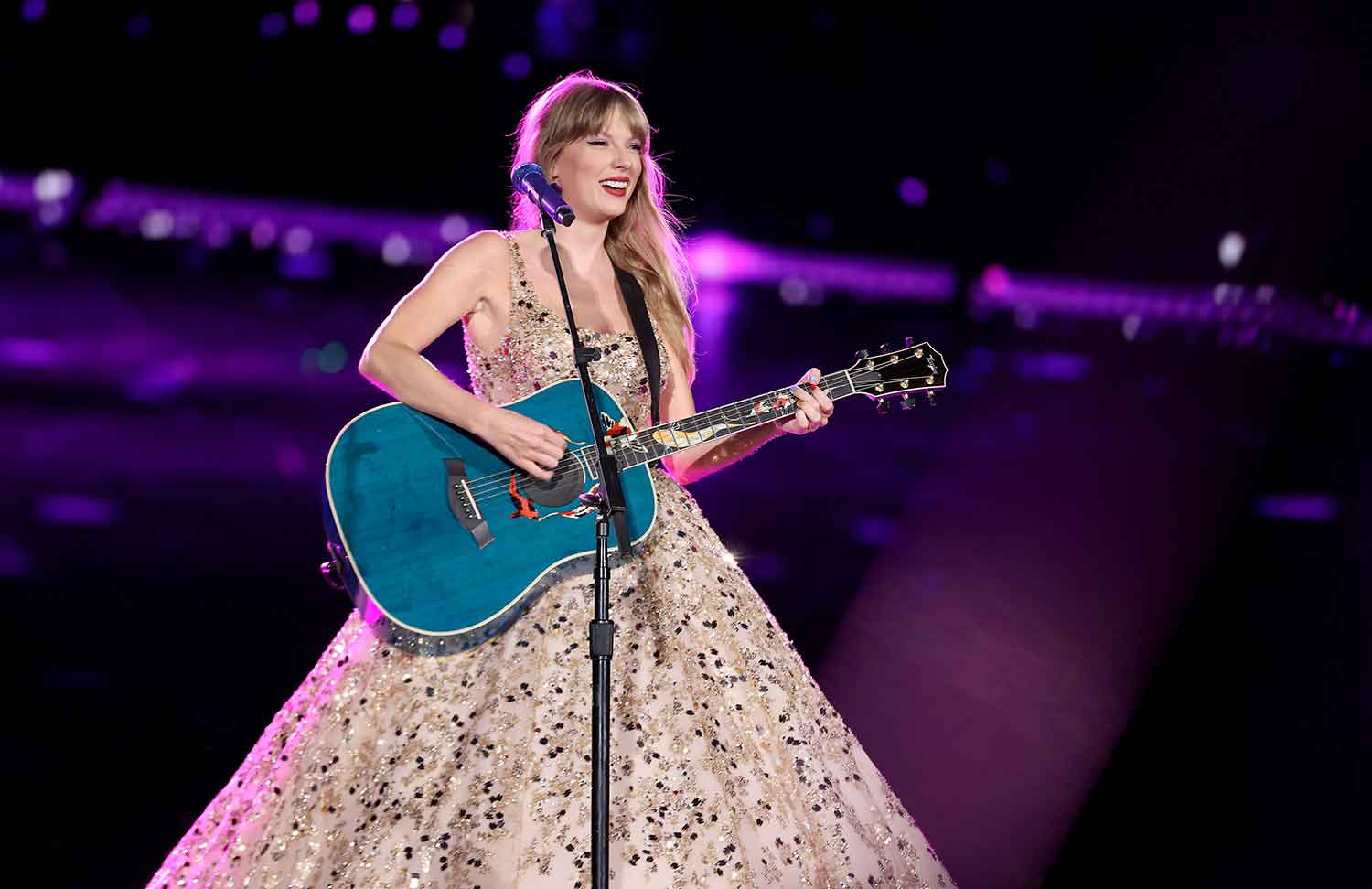 Taylor Swift holds a guitar and smiles while standing in front of a microphone and wearing a sparkly dress.
