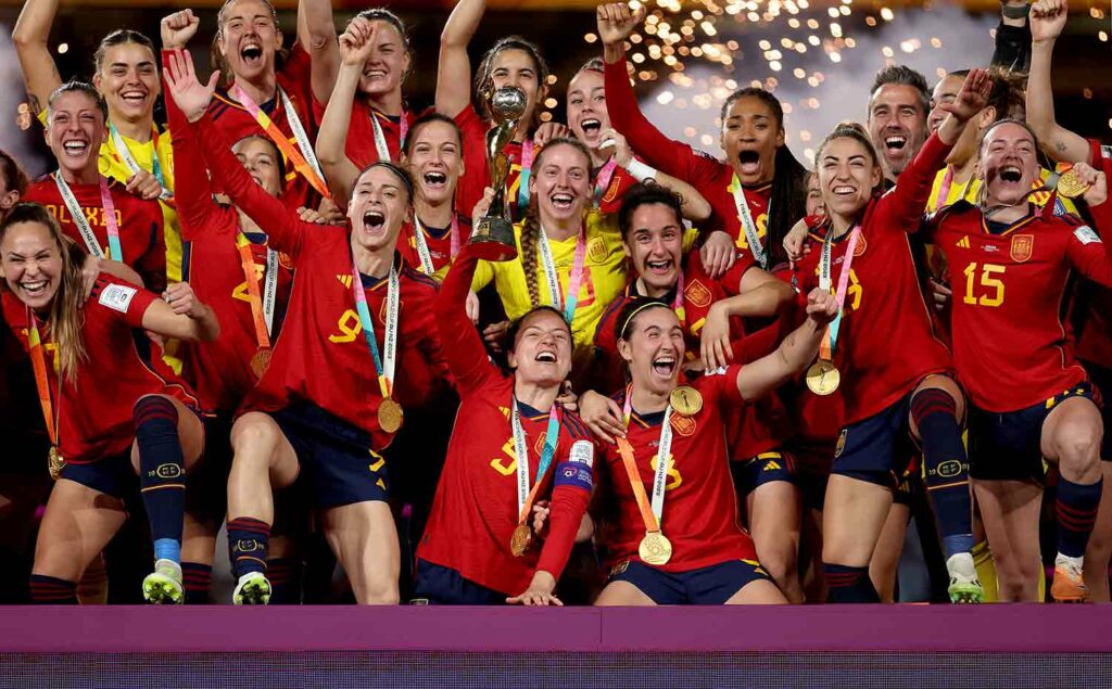 Women in red, yellow, and black uniforms pose and cheer with their arms up as one woman holds a trophy.