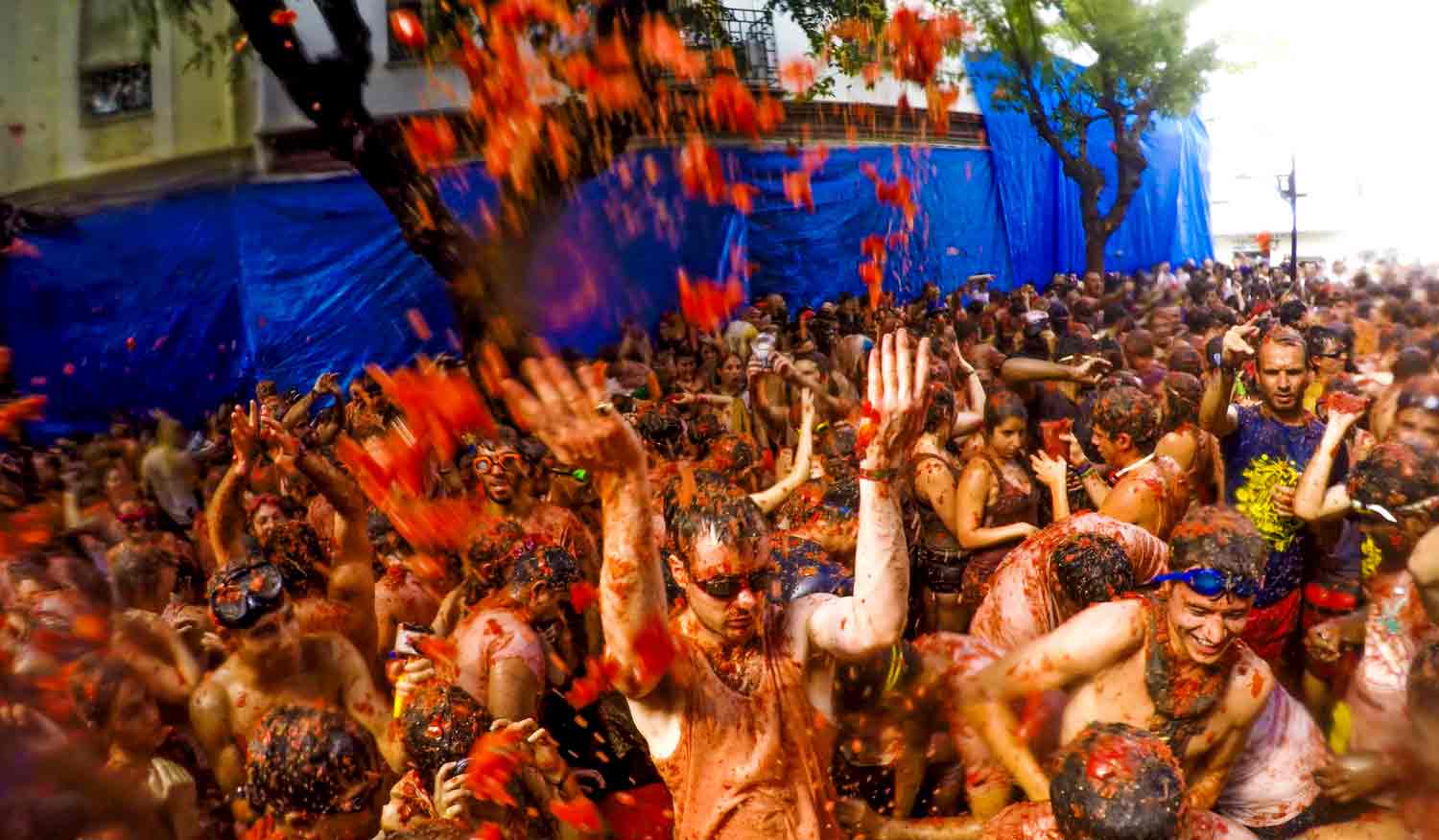 A man wearing goggles in the center of a tomato covered crowd throws crushed tomatoes into the air.