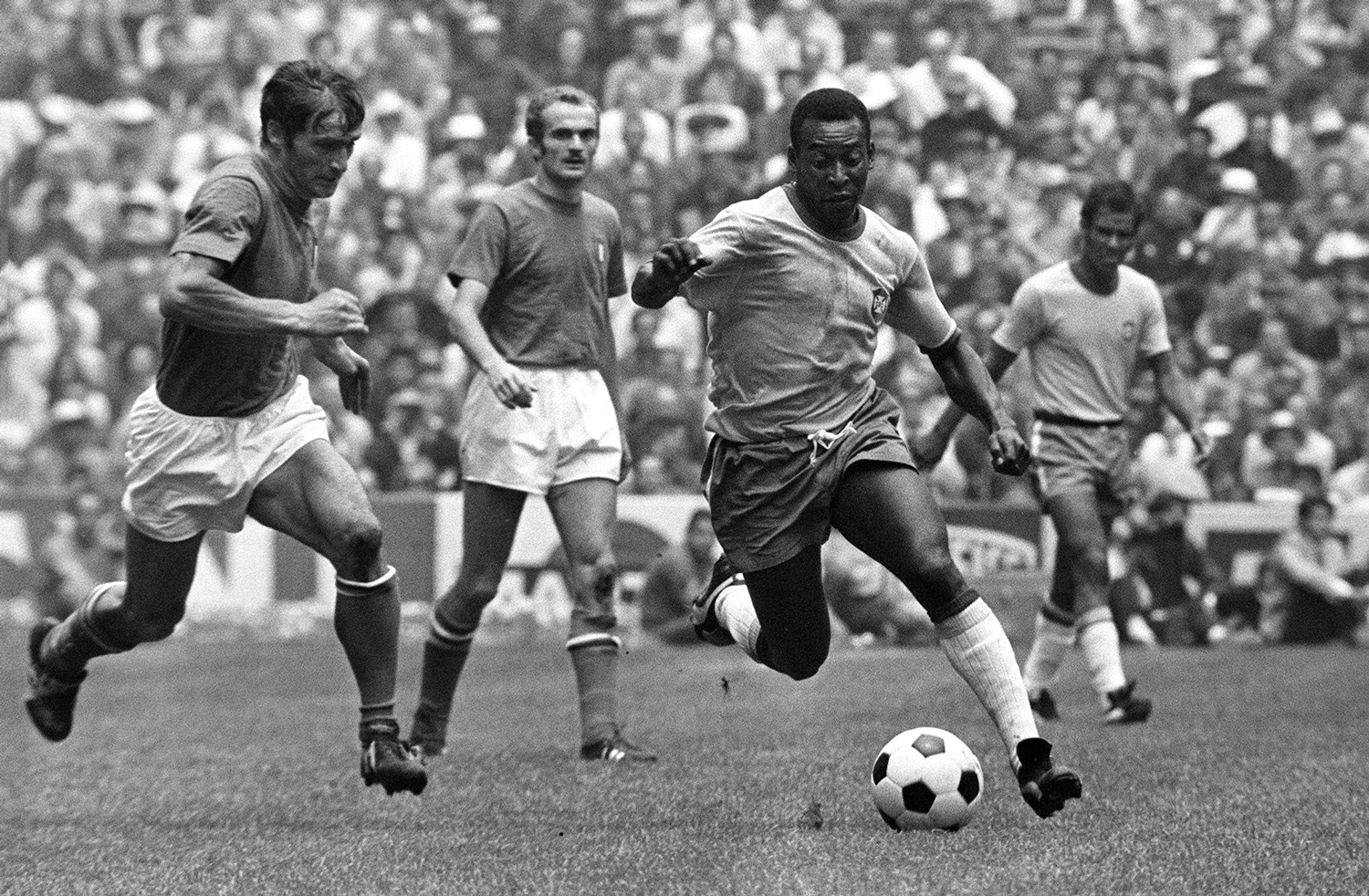 A man on a soccer field with three other players behind him is about to kick a ball