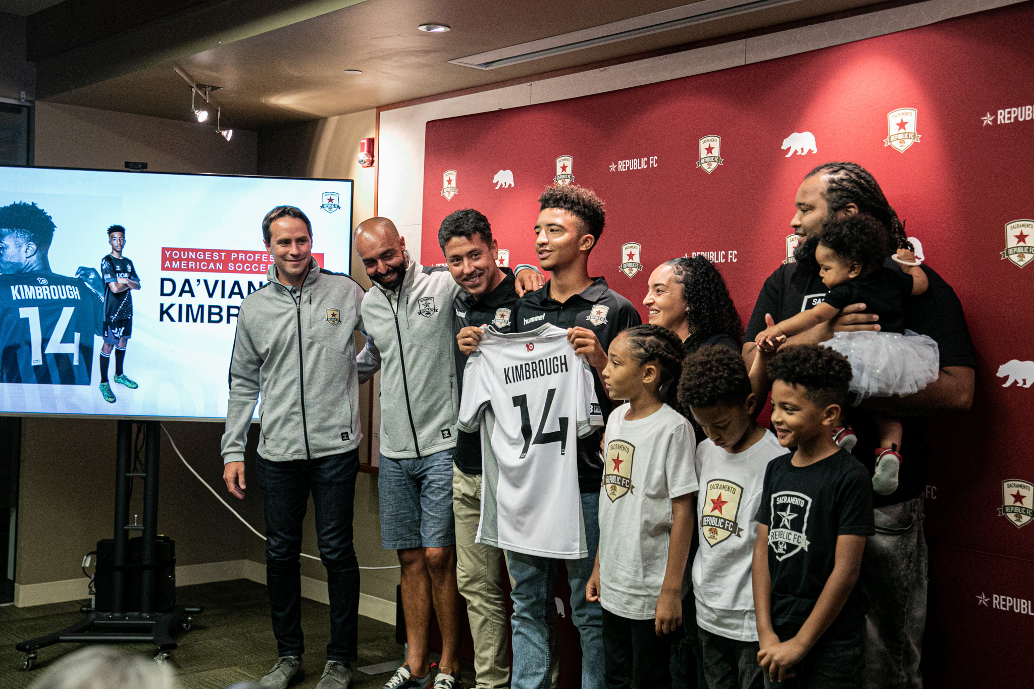 A teen holds up a white jersey reading Kimbrough surrounded by adults and children in front of a backdrop reading Republic FC.