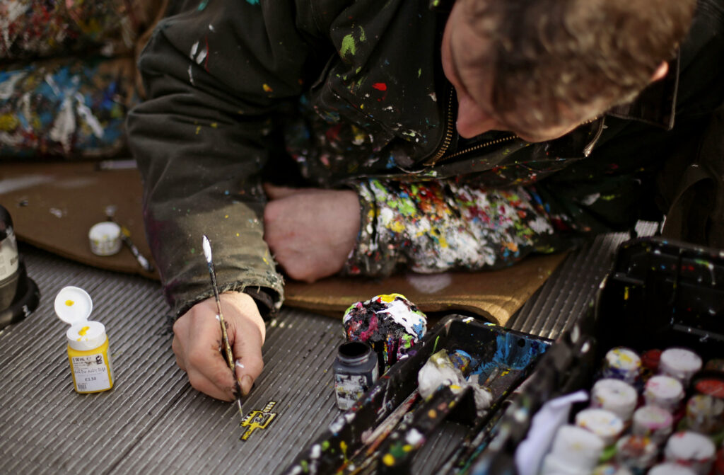 A man wearing clothes splattered with paint lies on his stomach and paints a design on something that is on the ground.