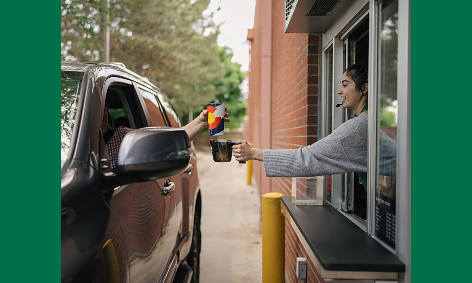 A worker at a drive-thru window holds out a beverage in a mug while the driver holds out another beverage container.