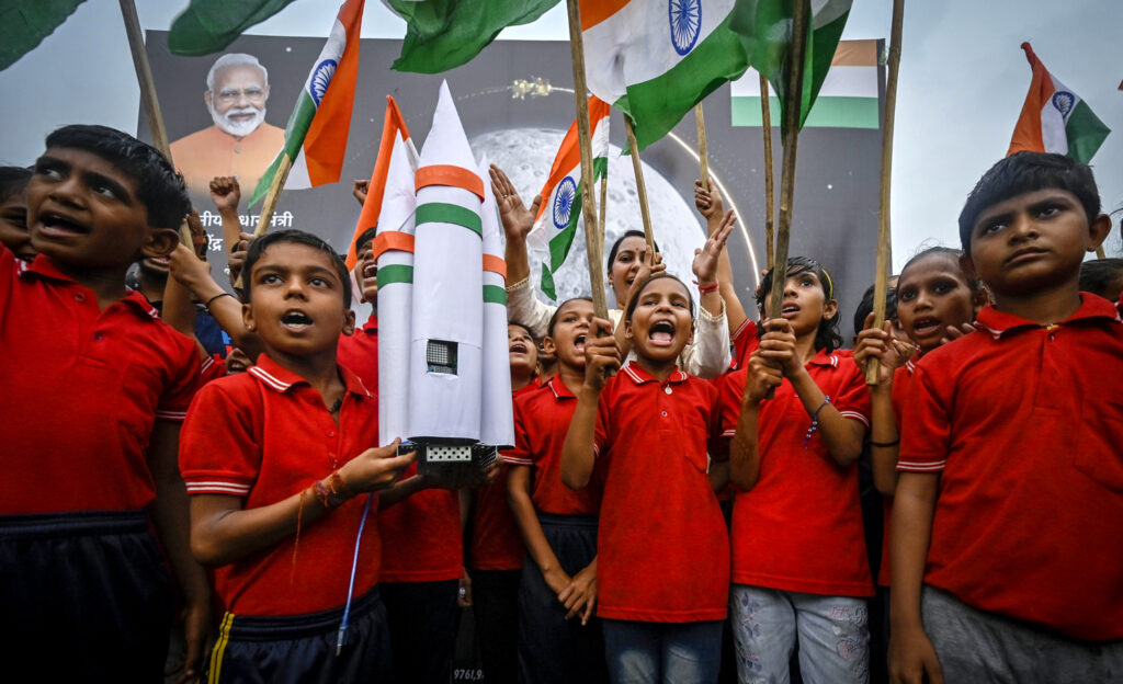 Children in red polo shirts wave Indian flags as one child holds a model of a spacecraft.