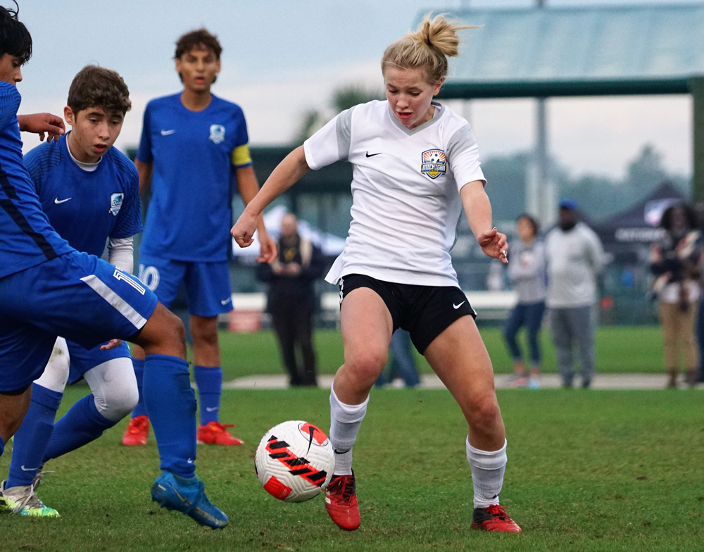 A teen in a white jersey and some teens in blue jerseys play soccer on a field.