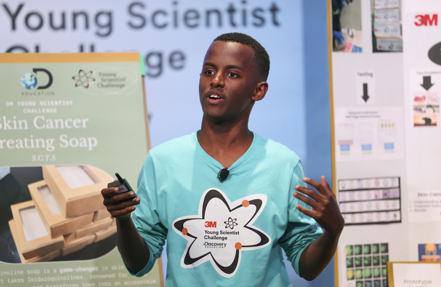 A teenage boy wears a 3M Young Scientist Challenge shirt and speaks in front of a display that says skin cancer treating soap.