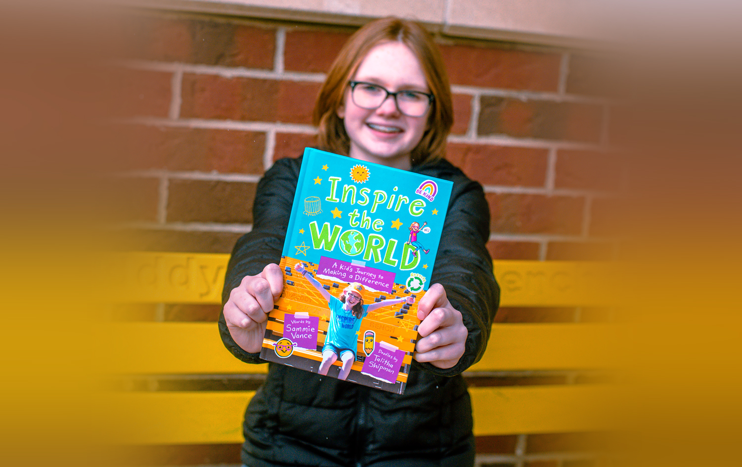 A teenage girl stands in front of a yellow buddy bench and holds out a book with the title Inspire the World.