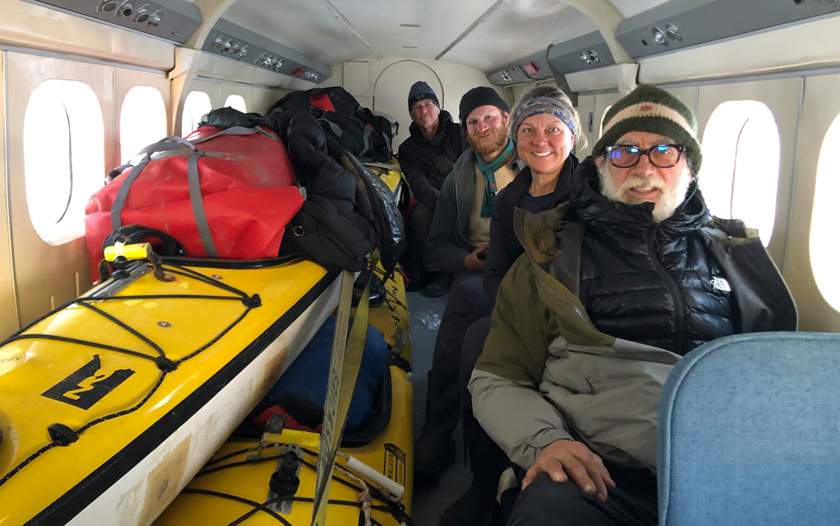 Three men and a woman sit inside a small airplane cabin alongside two kayaks.