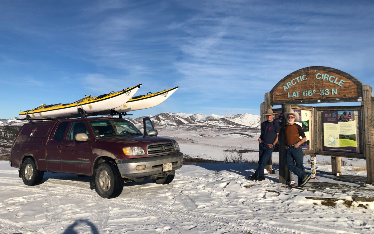 Two men in cowboy hats pose next to a sign that says Arctic Circle alongside an SUV with two kayaks strapped to its roof.