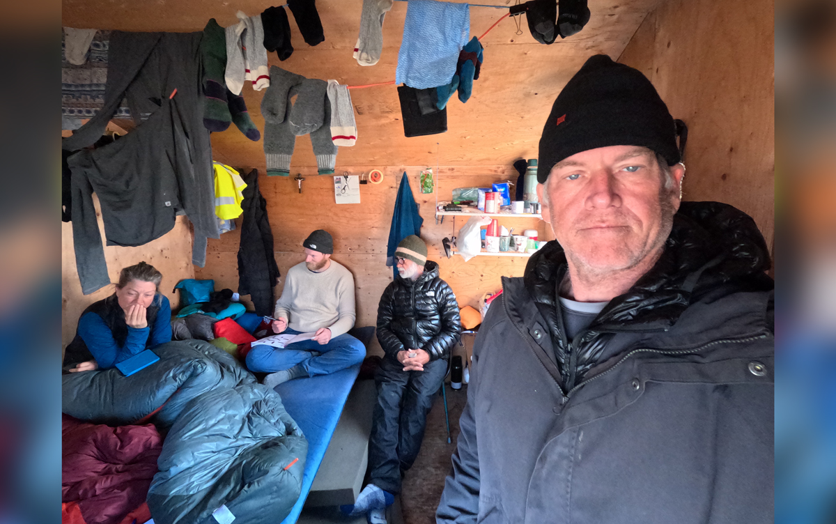 Three men and one woman wearing warm clothing inside a cabin with clothes hanging on lines.