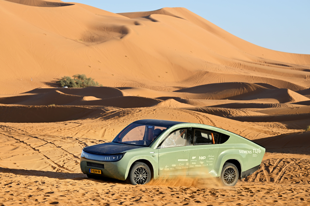 A green car drives across a desert landscape with many sand dunes.