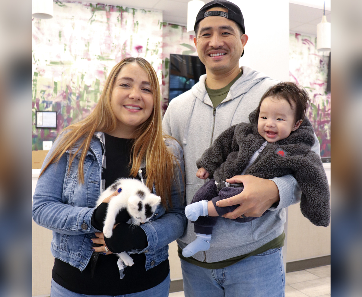 A man and woman pose for a photo. The man holds a smiling baby and the woman holds a kitten.