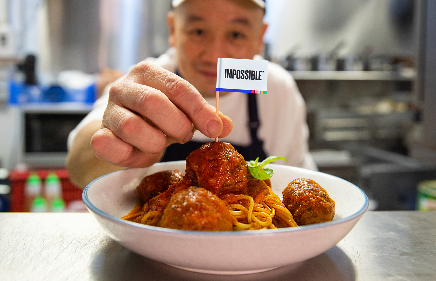 A man places a flag reading Impossible in a meatball that is part of a bowl of spaghetti and meatballs.