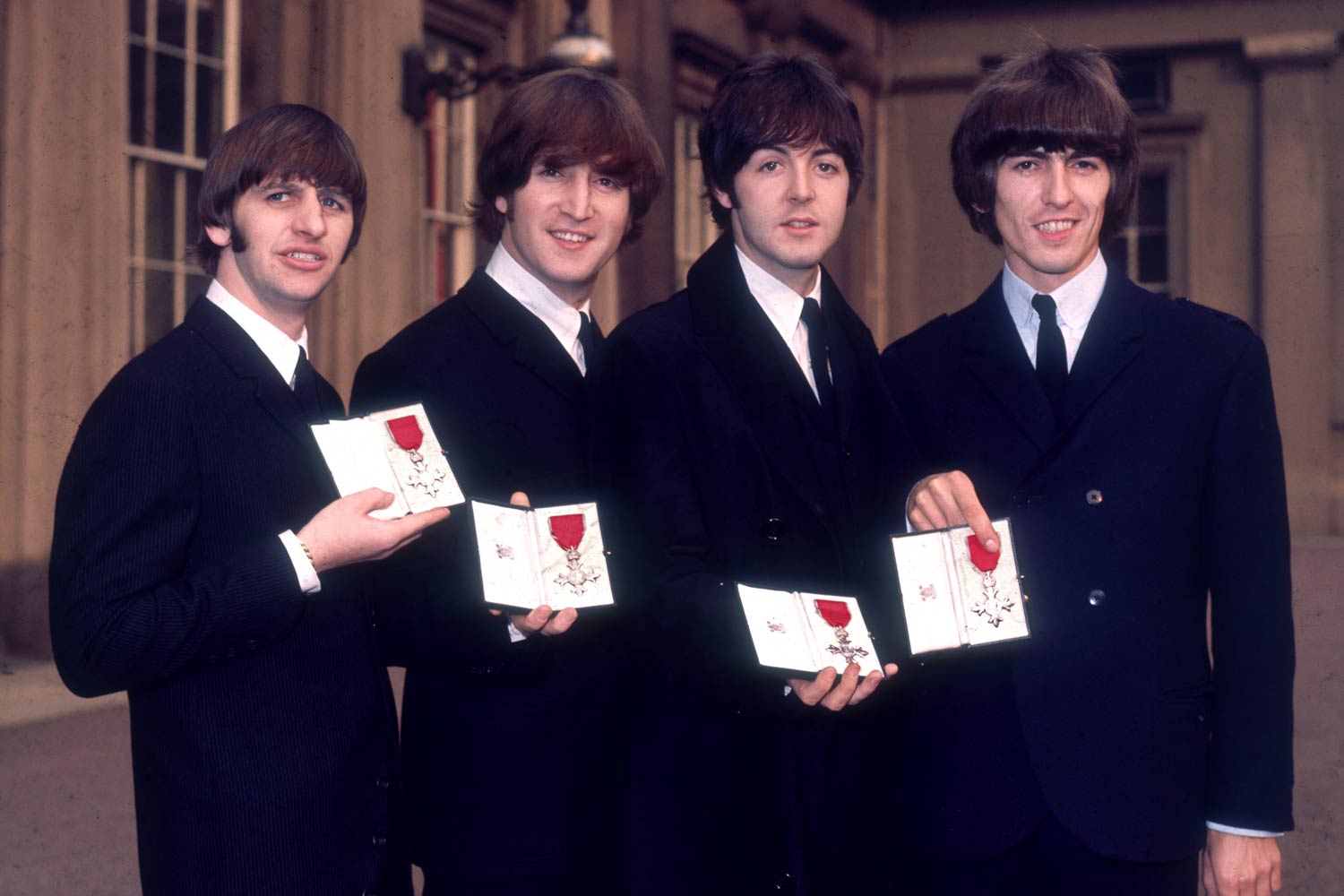 The Beatles in dark suits and black ties pose together, each one holding a medal in a small box.