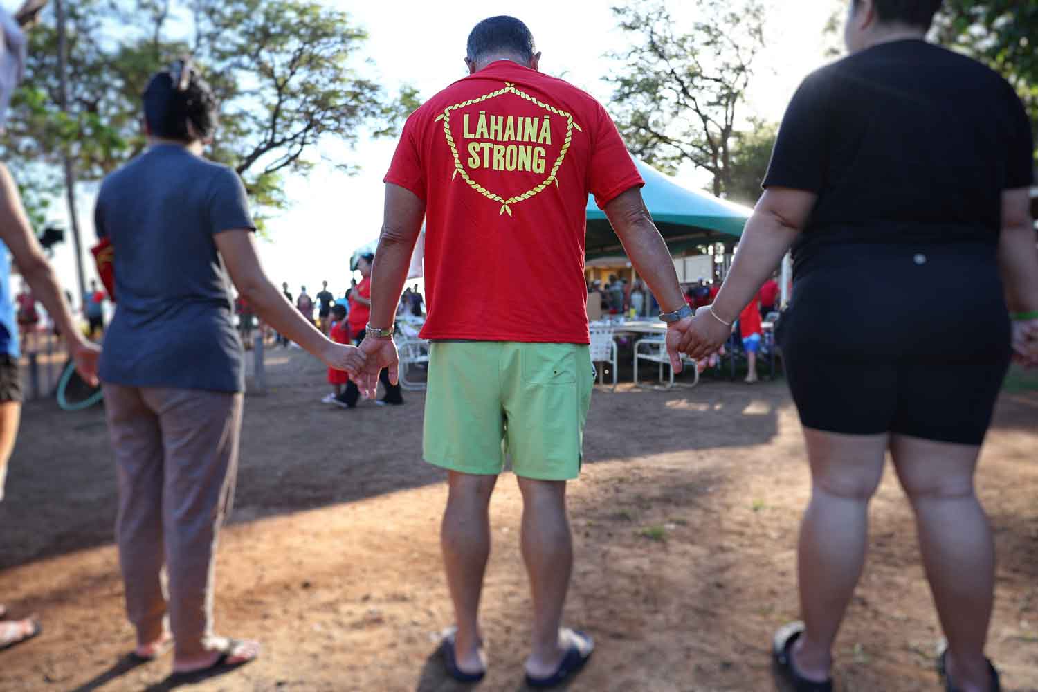 View from behind of three people standing next to each other and holding hands, with the man in the middle wearing a shirt that reads Lahaina Strong.