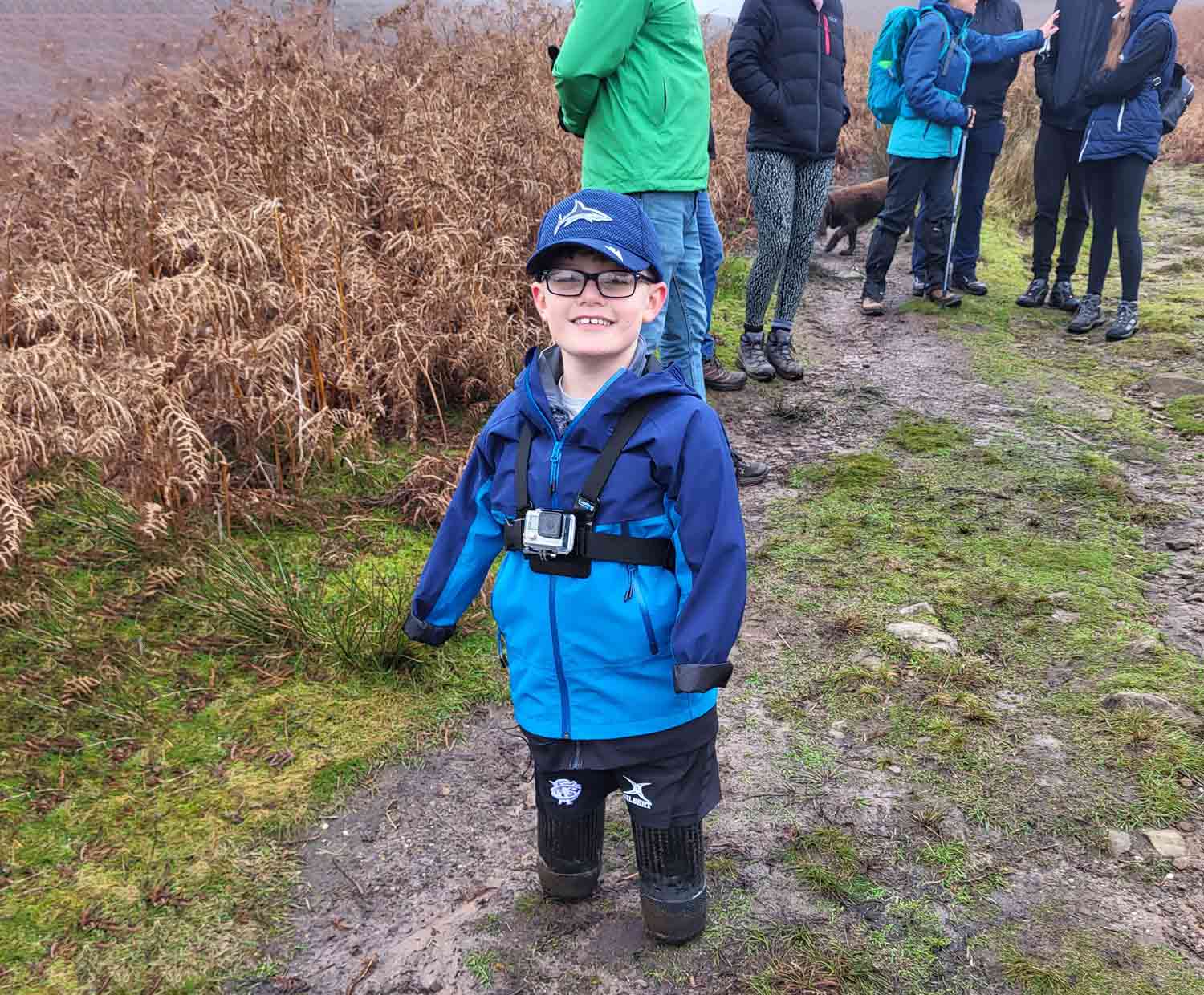 Luke Mortimer stands on a hiking trail and poses for the camera as several adults in hiking clothes can be seen behind him.