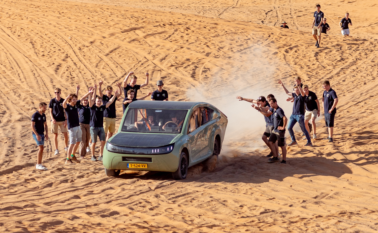 On a desert landscape, a crowd of people in blue shirts cheers as two people drive a small green car.