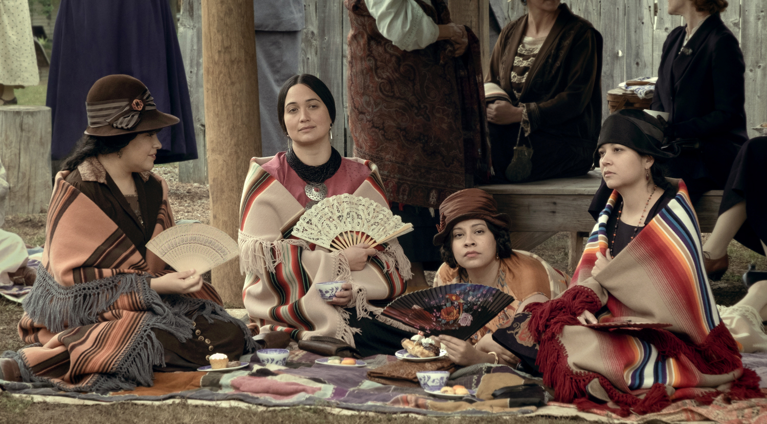 Lily Gladstone and three other actresses sit on a blanket wearing shawls and holding fans in a still from Killers of the Flower Moon.