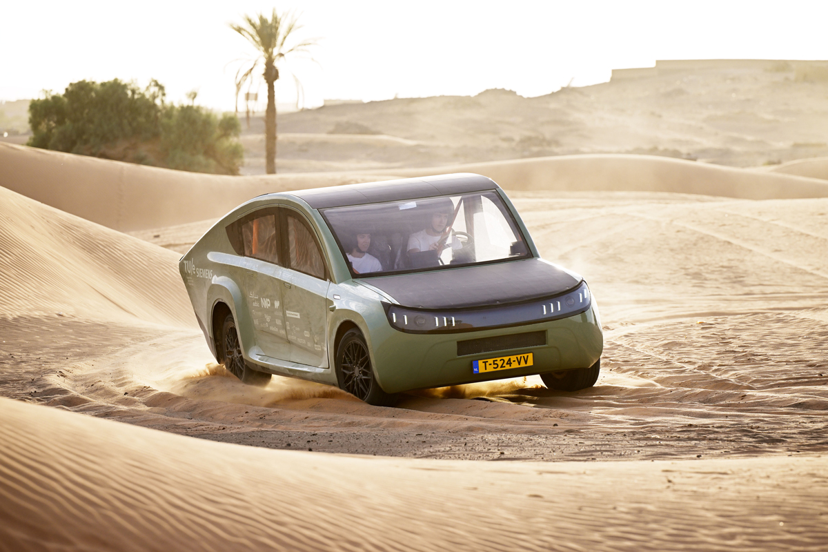 Two people wear helmets as they drive a green car on sand.