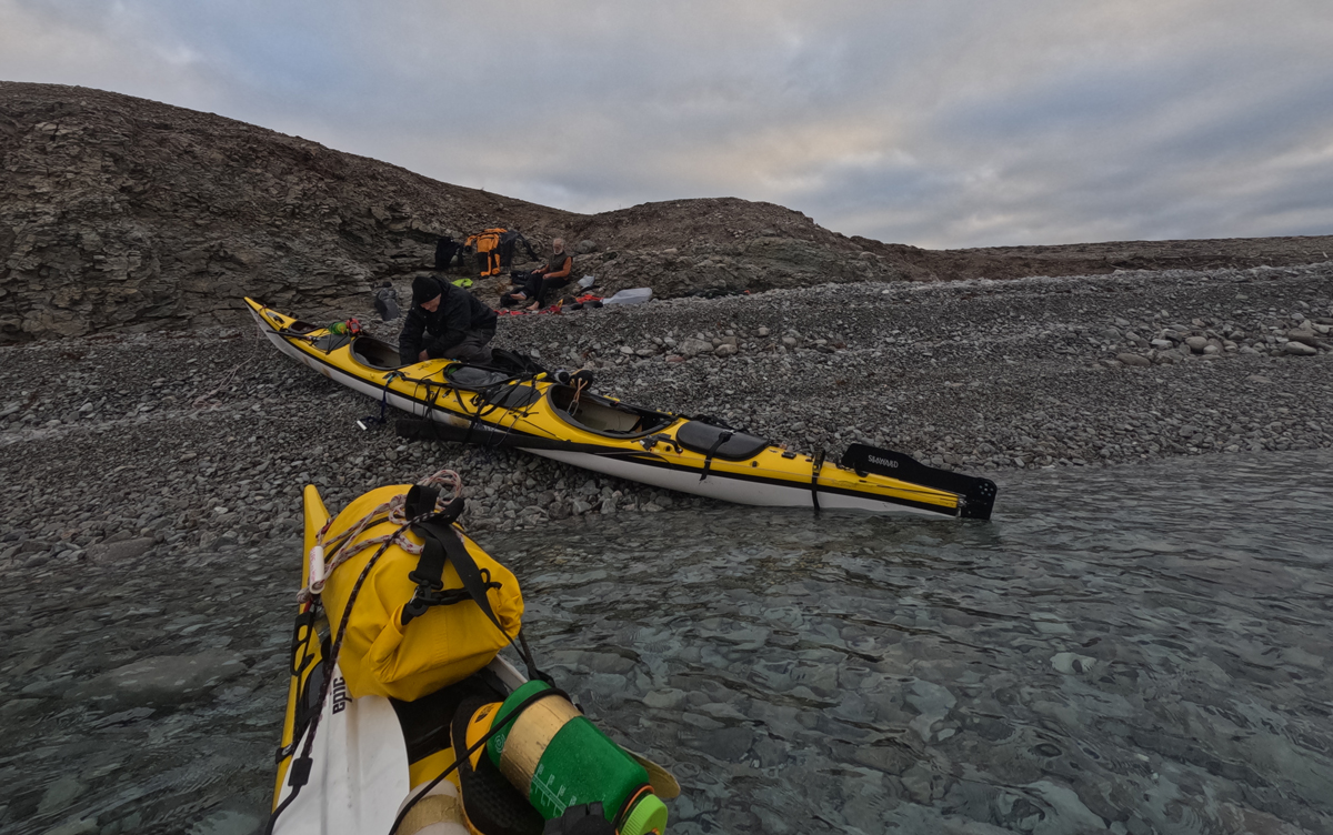 A man sits near a yellow kayak on a rocky shore, and another kayak can be seen.