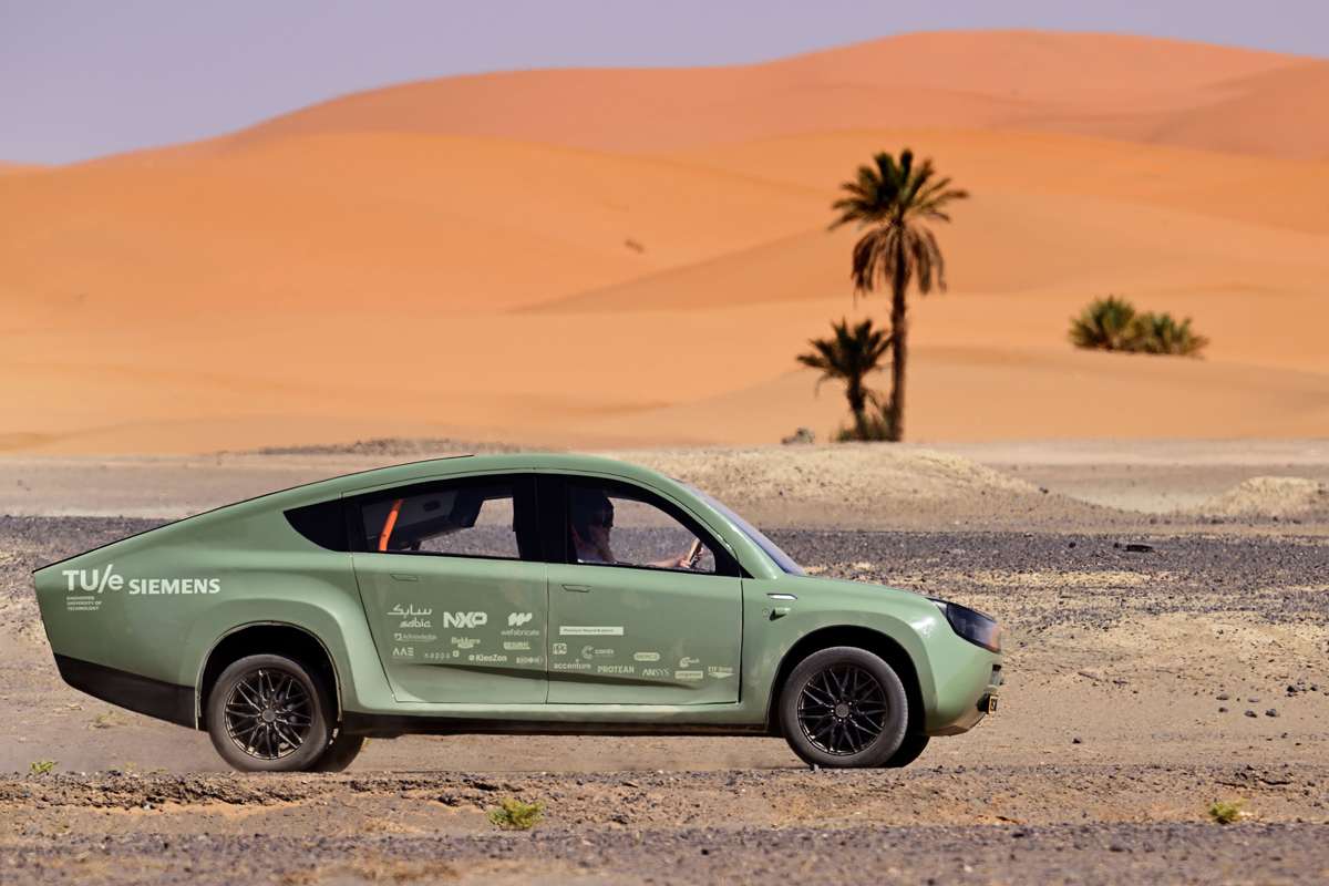 A side view of a green car bearing company logos being driven in a desert.