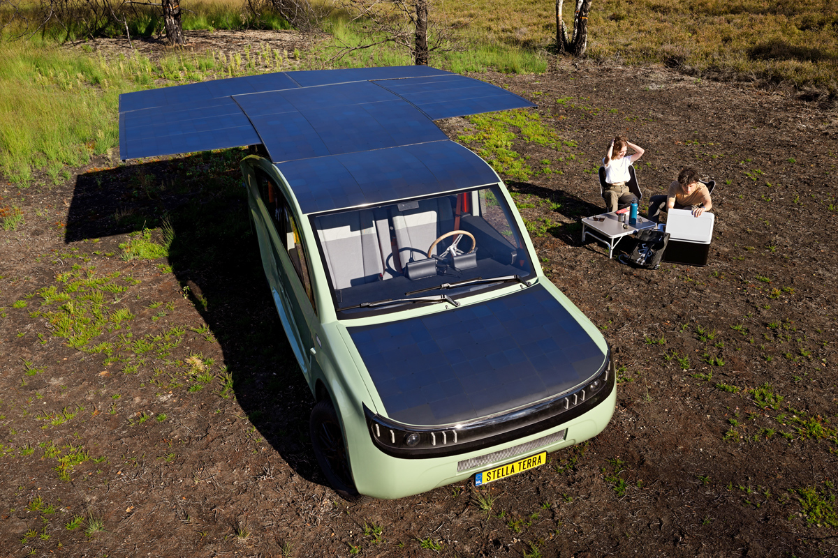 Two people sit at a table next to a car with large solar panels extending from its roof.