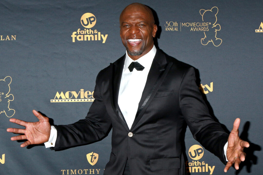 Terry Crews smiles and poses in front of a backdrop reading MovieGuide Awards with arms outstretched and wearing a tuxedo.