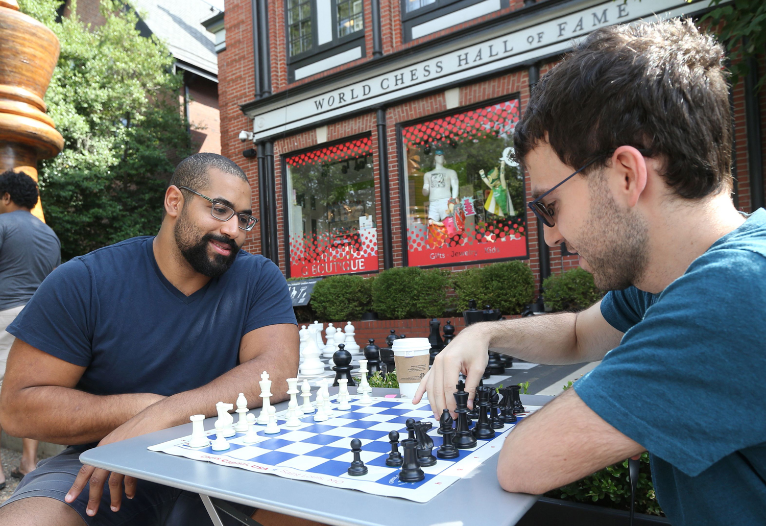 John Urschel and another man sit at an outdoor table and play chess in front of a storefront with a sign reading World Chess Hall of Fame.