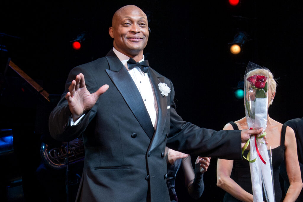 Eddie George stands on stage wearing a tuxedo, holding flowers, and smiling.
